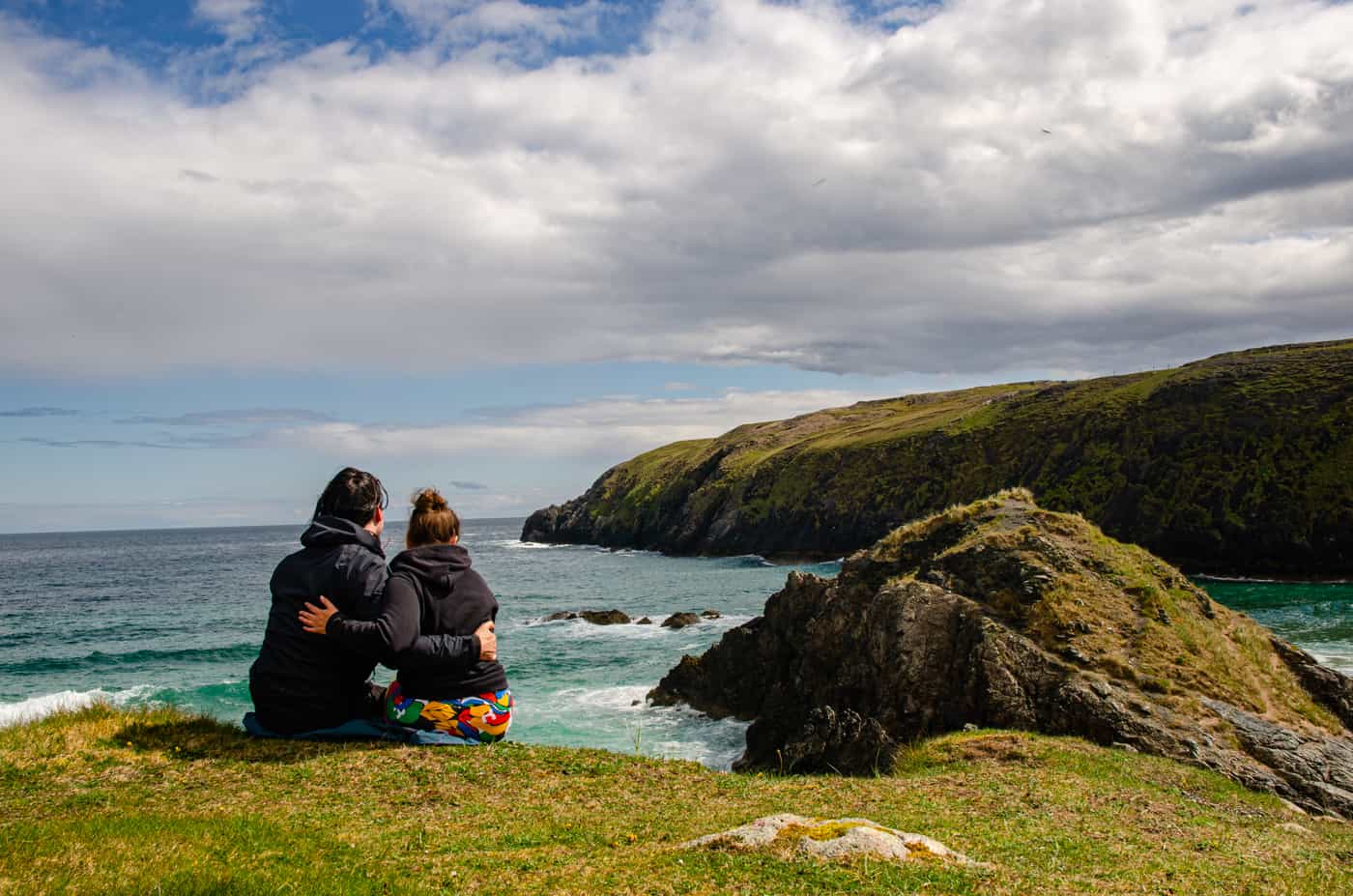 A couple sitting on a cliff watching sea and beach in summer in Scotland.
