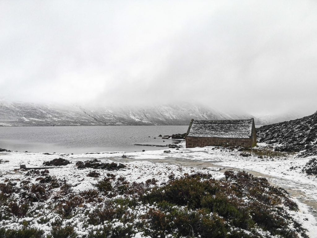 View at snow-covered landscape with an old cottage, lake and mountains in a fog. Cairngorm is one of the best places to visit in UK in February.