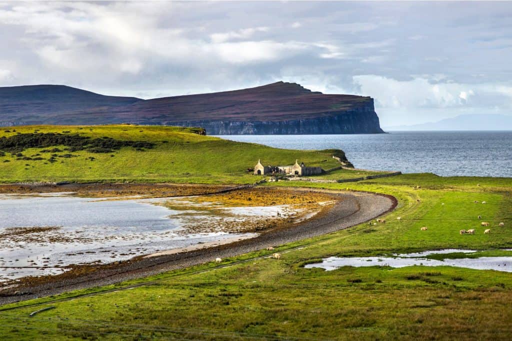 A view at meadow with sheep, lake, old cottage, sea and rock formation in distance.