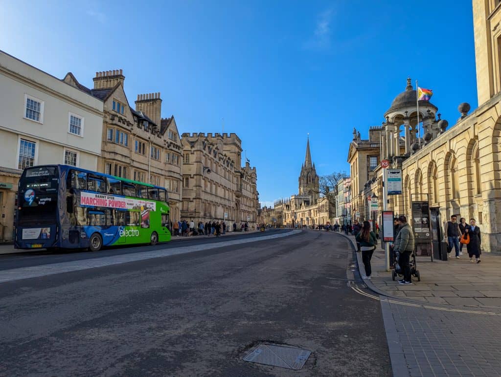Busy street in Oxford with people, mediaeval buildings, a bus and people.