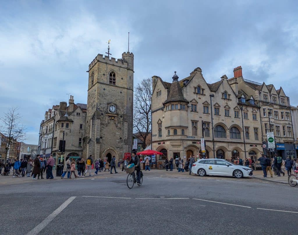 Busy road in Oxford with people, mediaeval tower and buildings at back.