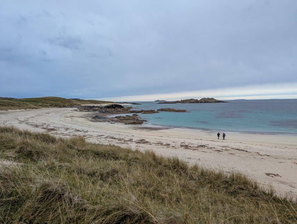 A beach on Iona island with people walking on a long beach on a cloudy day. Iona Island has one of the best Isle of Mull walks.