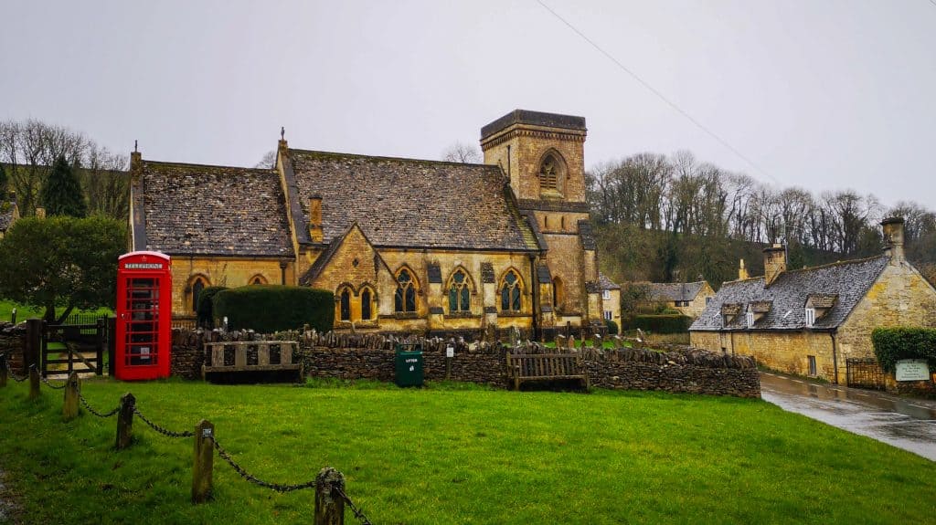 A sand-coloured old church and cottage with a red telephone box in Snowshill Costwolds on a rainy day